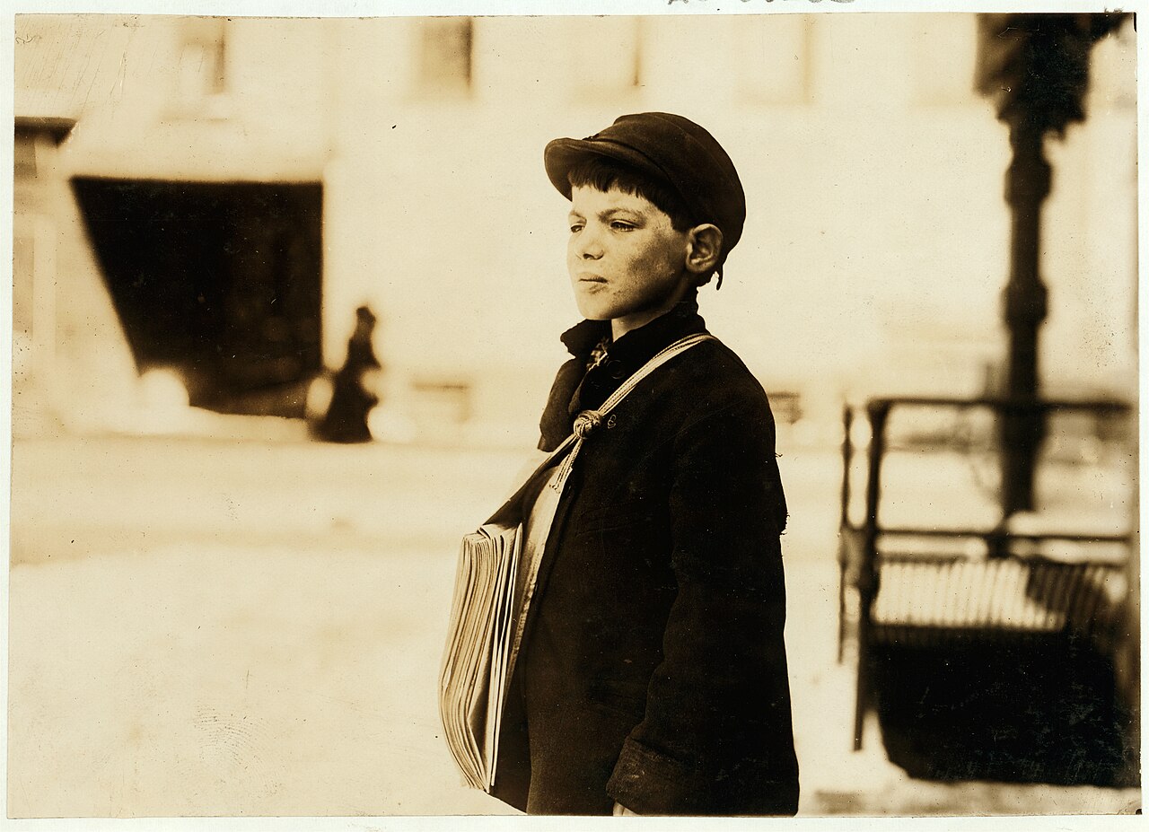 A newsboy on a street corner, stack of papers under his arm, shouting a headline.