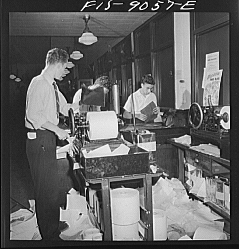 A wall of teletype machines in a 1950s wire service office, operators tending multiple feeds.