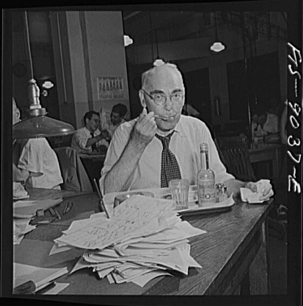An editor at a desk, red pencil in hand, marking up galley proofs spread across the surface.
